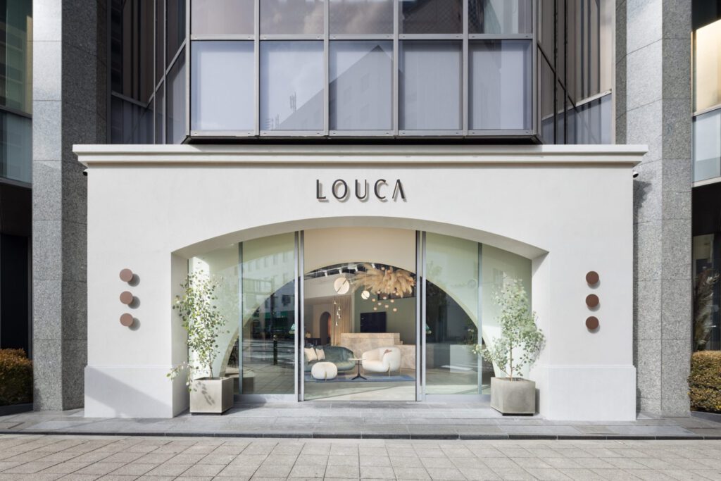 インテリアショップ LOUCA の路面の外観。白い壁のアーチ形の開口にガラスの扉、ガラス越しに店内のソファと照明が見え、入口の両脇に鉢植えの木（インフィニティ株式会社）