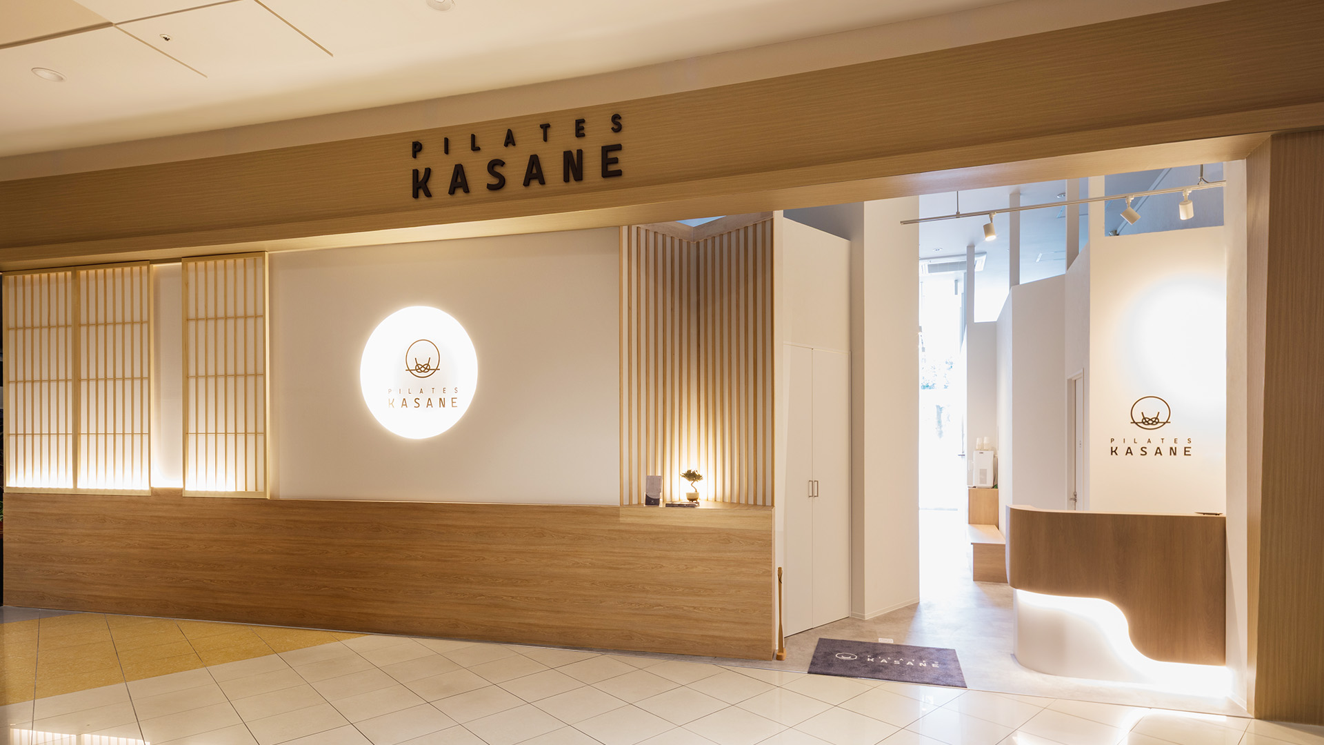 千葉県のピラティススタジオの施工写真：商業施設内のファサード、木ルーバーと発光ロゴサイン、曲面の受付カウンター｜施工：カエル・デザイン・プロジェクト株式会社