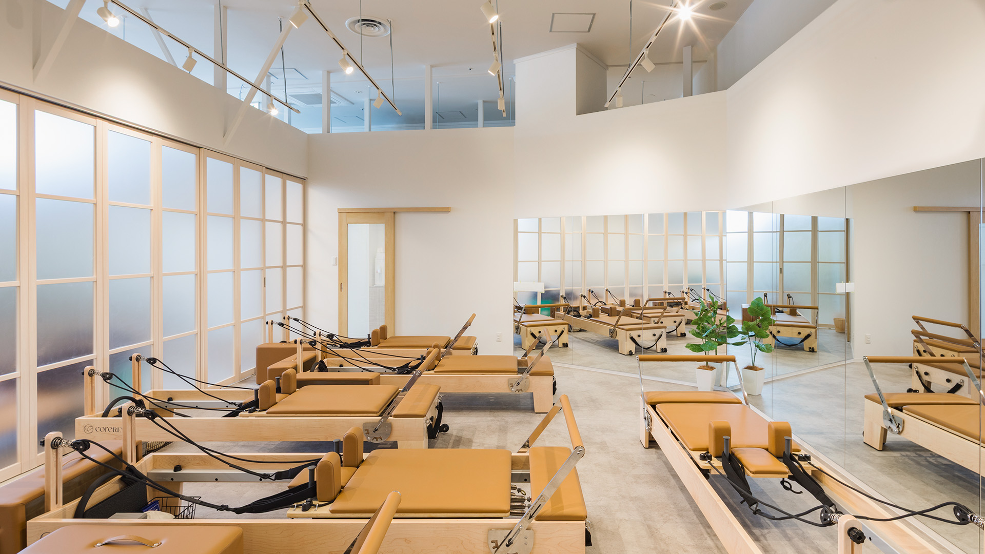 千葉県のピラティススタジオの施工写真：吹き抜けの窓面に木製リフォーマーを2列配置したマシンピラティスのスタジオ、正面は全面鏡｜施工：カエル・デザイン・プロジェクト株式会社