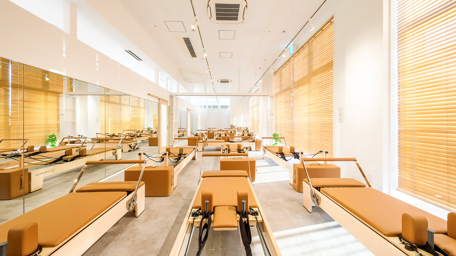 沖縄県のピラティススタジオの施工写真：木製ブラインドの窓側にリフォーマーを並べたマシンピラティスのスタジオと壁一面の鏡｜施工：カエル・デザイン・プロジェクト株式会社