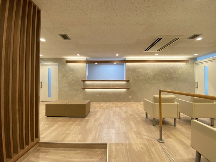多摩市のクリニック 東京都多摩市 歯科クリニック 木目×グレー 待合。木目の長尺シートの床とベージュのソファ、塗り壁風のグレーの壁、天井ラインの木製間接照明（株式会社S plus Design’s）