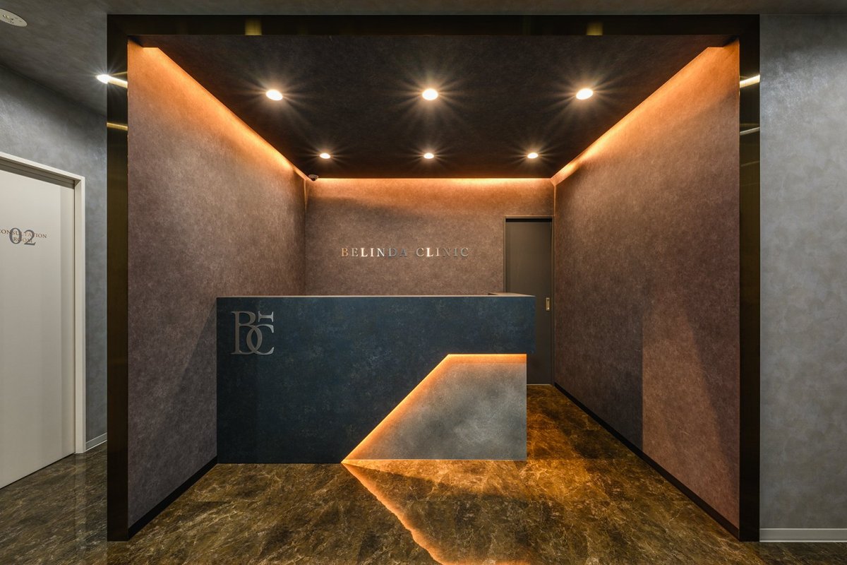 BELINDACLINIC 柏院 千葉県 美容クリニック ダークグレー×間接照明・ラグジュアリー 受付。ダークグレーの壁と光るカウンター、オレンジの間接照明(サンリット建設(DESIGN STUDIO TEMPO))