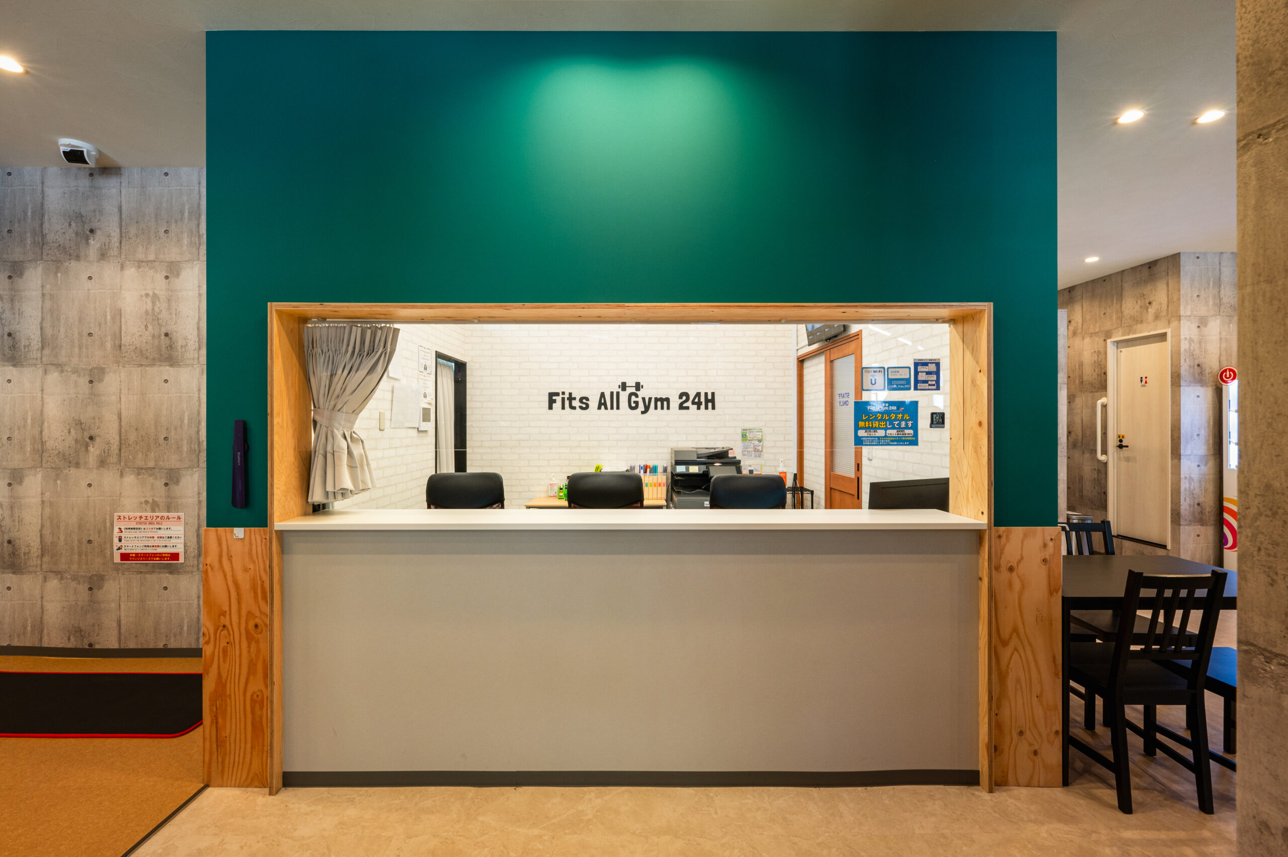 Fits All Gym 24H 市沢町店の受付。緑の壁に木の枠の受付窓とカウンター、奥の白いれんが調の壁に店名のロゴ。施工：株式会社サンリット建設