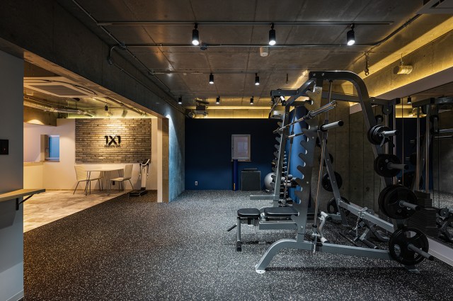 PERSONAL GYM 1×1 のトレーニングエリア。コンクリートの天井にスポットライト、紺の壁、白い粒の入った黒いゴムの床に複数のラック。奥にロゴの壁とテーブル席（株式会社サンリット建設）