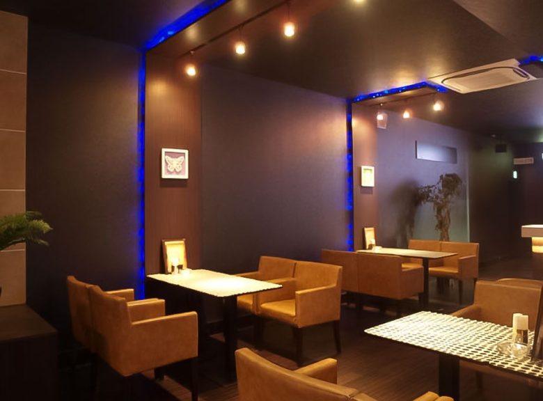 ダーツバーのソファ席。茶色の一人掛けソファとテーブル、天井と柱の縁に青いライン照明、天井から下げた照明（タカハシアートプランニング株式会社）