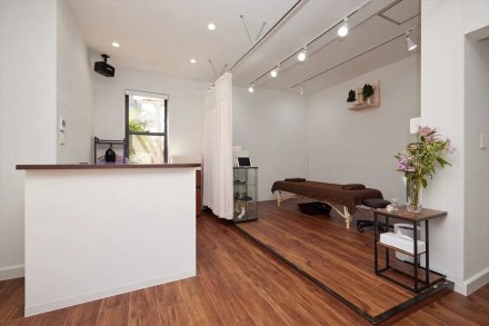 寝屋川あおば腰痛整体院（大阪府寝屋川市）の受付と施術スペース。手前に白い受付カウンター、一段上げた木目の床に施術ベッド、天井のレールの白いカーテンとスポットライト（ユウリフォーム）