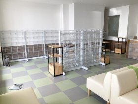 あいり薬局（東京都足立区）の待合。白いワイヤーの陳列棚と木の小さな棚、緑のラインのソファ、市松の床（施工：株式会社ビリーフデザイン）