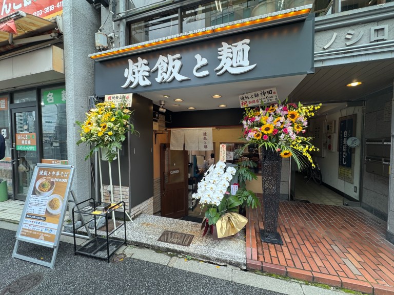 焼飯と麺 神田の外観：濃い灰色の壁に白い字の看板、開店の花、メニューの立て看板、れんが色のタイルの床