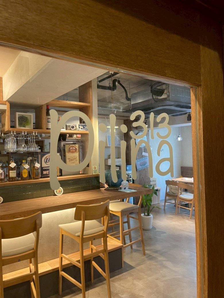 Pilina313 神戸市元町商店街 クラフトビールバー。木枠の開口越しに見える木のカウンターとハイチェア、背面棚のボトル、ガラスに店名ロゴ、天井は現し（LEBON DESIGN）