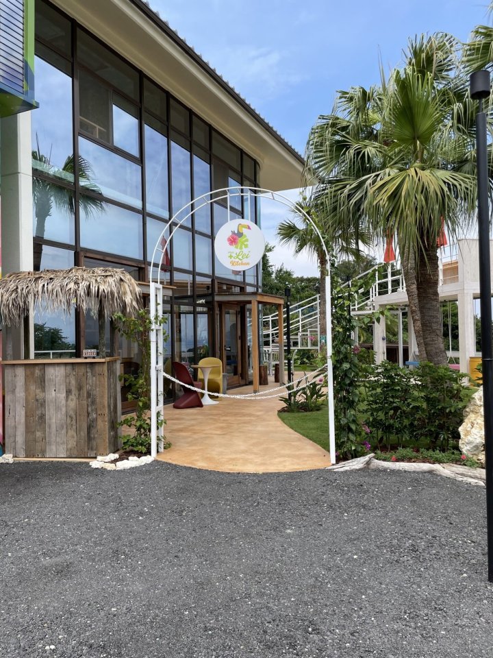 Plus Lei Kitchen 淡路市 飲食店の外構。大きなガラス窓の建物とヤシの木、白いアーチのゲート、ヤシ葺き屋根の小屋があるハワイアンリゾート風のアプローチ（LEBON DESIGN）