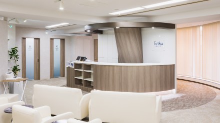 整痛院ふっか 銀座（東京都中央区）の待合と受付。中央に木目のラウンド型の受付カウンター、手前に白い1人掛けのソファ、左奥にすりガラスの扉の部屋が並び、右は縦型のブラインドの曲面の窓（カエル・デザイン・プロジェクト株式会社）