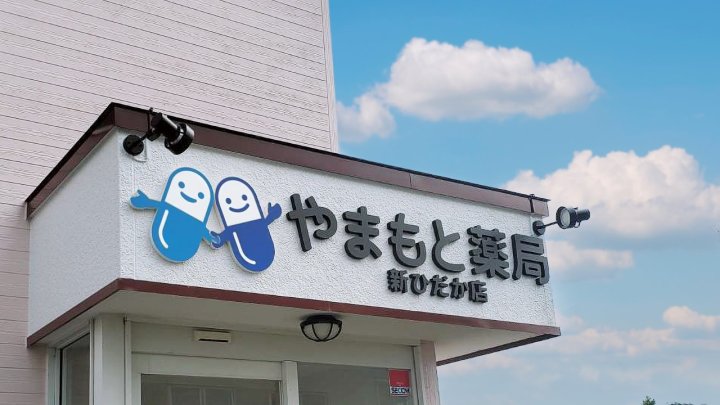 やまもと薬局 新ひだか店（北海道日高郡）の入口上の看板。白い看板面に青いカプセルのキャラクターと黒い切り文字の店名、スポットライト（施工：カエル・デザイン・プロジェクト株式会社）