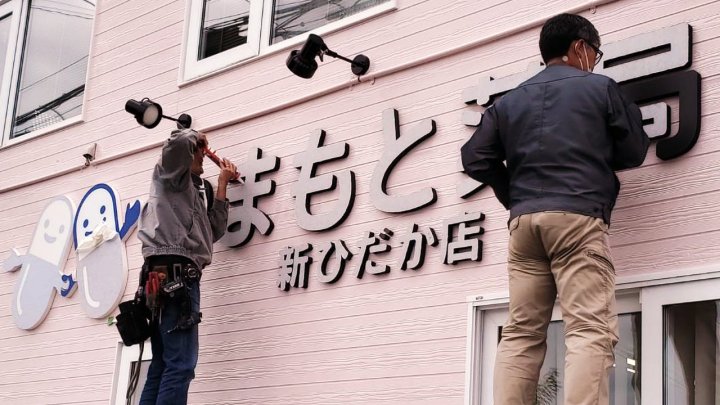やまもと薬局 新ひだか店（北海道日高郡）のサイン施工中。外壁に「やまもと薬局 新ひだか店」の切り文字サインを取り付ける作業（施工：カエル・デザイン・プロジェクト株式会社）