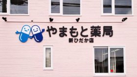 やまもと薬局 新ひだか店（北海道日高郡）の外観。ピンク系の外壁にキャラクター入り「やまもと薬局 新ひだか店」のサイン（施工：カエル・デザイン・プロジェクト株式会社）