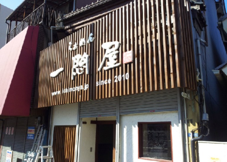 らぁめん 一悶屋の外観：2階の前を縦に並べた木の板で覆い、白い字の店名を付けた看板、白い壁の1階の入口
