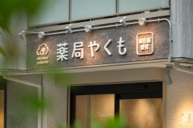 薬局やくも（東京都・都立大学）の外観サイン。グレーの看板帯に白い文字の店名と「処方箋受付」の表示、アームライト（施工：株式会社サンリット建設）