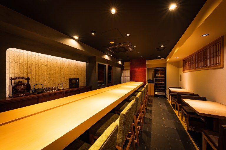 銀座創作中華（東京都・銀座）の店内。白木の長いカウンターに布張りの椅子が並び、奥の壁は灯りで照らした模様のタイルの棚に中国茶の道具を飾る。右に格子の窓とテーブル席（デザイン・施工：株式会社サンリット建設〔DESIGN STUDIO TEMPO〕）