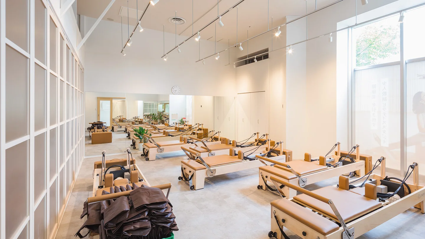 Pilates KASANE イオンタウンおゆみ野店（千葉市・ピラティススタジオ）。高い天井と大きな窓があり、木製のピラティスマシンを並べたスタジオ。