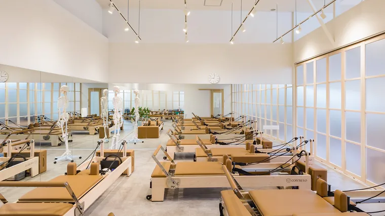 Pilates KASANE イオンタウンおゆみ野店（千葉市・ピラティススタジオ）。鏡張りの壁と格子状の間仕切りの間に、ピラティスマシンを並べています。