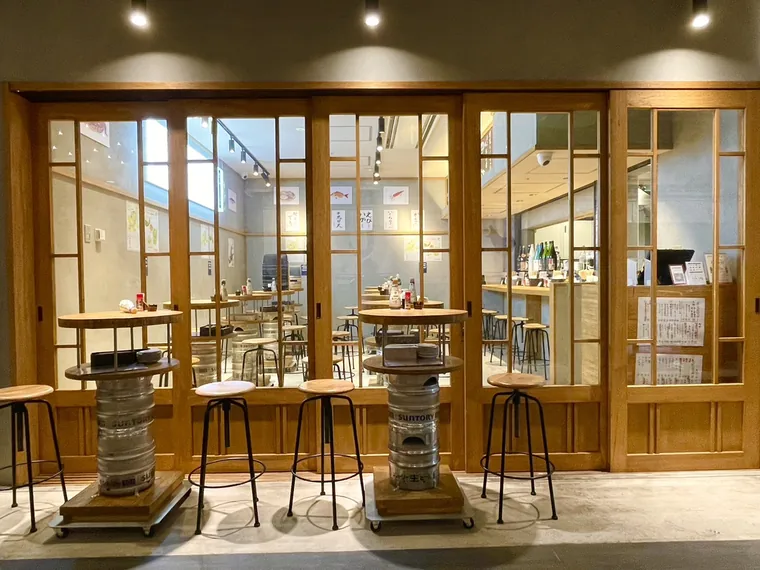 ニカイノオアシス KITTE大阪店（大阪府大阪市北区・飲食店）のファサード・客席。木製の建具とガラス越しに店内が見えます。通路側には丸いテーブルと椅子を置いています。
