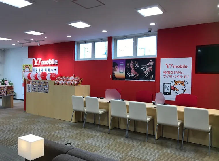 ソフトバンクワイモバイル 入間中央（入間市・携帯電話ショップ）。赤い壁の前に椅子と接客カウンターを並べた携帯電話ショップ。原典の工事範囲は店内ブースの新設です。